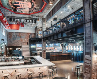 Spacious industrial-style sports bar interior with L-shaped marble bar, round stools, mezzanine seating, exposed brick walls, multiple TV screens and stage lighting