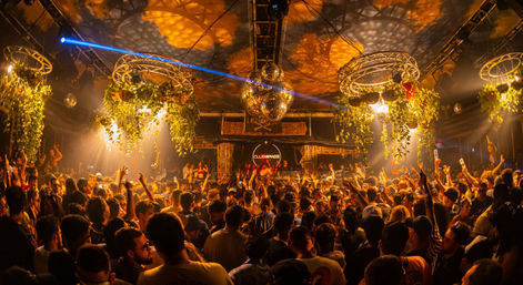 Packed indoor nightclub dance floor at a lively late-night party with warm amber lighting, disco ball, hanging greenery, laser beam, and DJ on stage.