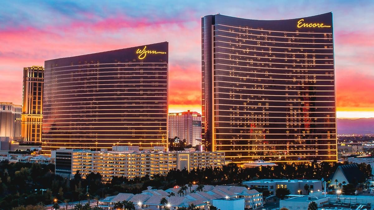 Sunset over two curved luxury resort towers on the Las Vegas Strip, glass façades glowing with horizontal gold lights and reflected neon against a vivid pink-orange sky.