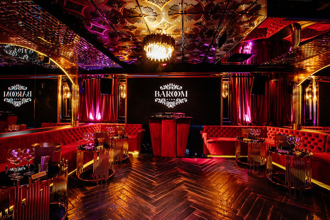 Luxurious nightclub lounge with red velvet banquettes, mirrored ceiling and chandelier, central DJ booth, purple-gold ambient lighting, and herringbone wood floor.