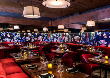 Cozy upscale restaurant interior with red velvet banquettes, floral backlit mural, hanging pendant lights and candlelit wooden tables set for dinner.
