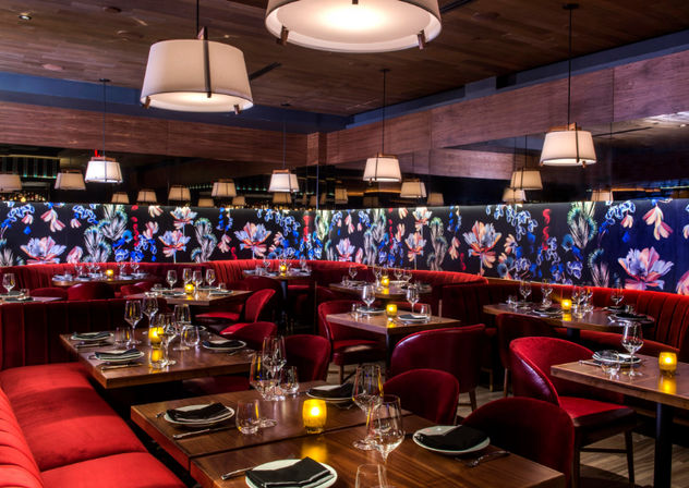 Cozy upscale restaurant interior with red velvet banquettes, floral backlit mural, hanging pendant lights and candlelit wooden tables set for dinner.