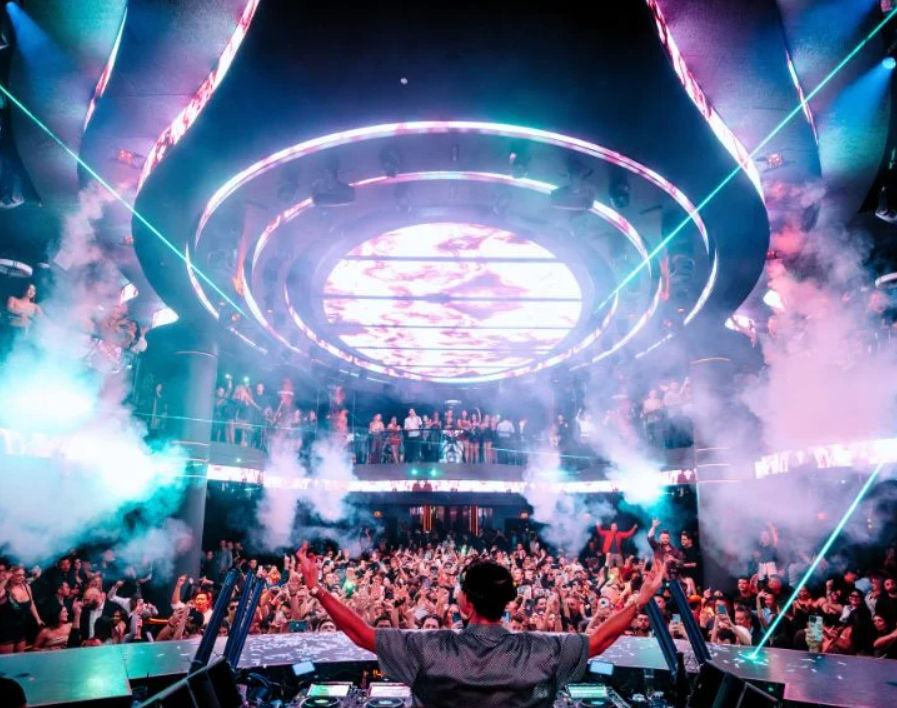 DJ with arms raised at a packed multi-level nightclub, neon circular LED ceiling, laser beams and smoke lighting up a dancing crowd