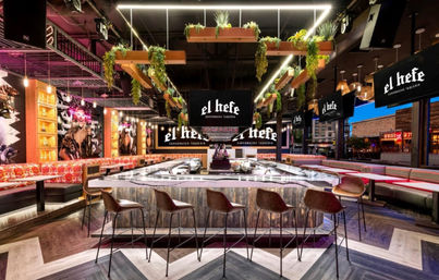 Vibrant modern Mexican restaurant interior with a central illuminated bar island, leather bar stools, hanging planters, neon-accent lighting, colorful booth seating and mural art, with a city street visible through large windows.