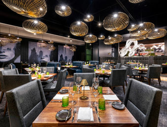 Contemporary upscale restaurant interior with wooden tables set for dining, gray upholstered chairs, green glassware, sculptural bar art and glowing gold wire pendant lights.