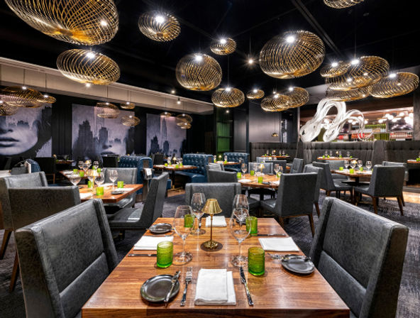Contemporary upscale restaurant interior with wooden tables set for dining, gray upholstered chairs, green glassware, sculptural bar art and glowing gold wire pendant lights.