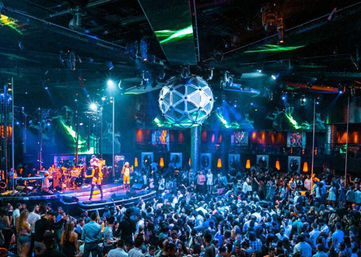 Energetic indoor nightclub scene with a live band on a lit stage, a large geometric disco ball overhead, colorful lasers and spotlights, and a packed dancing crowd.