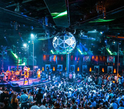 Energetic indoor nightclub scene with a live band on a lit stage, a large geometric disco ball overhead, colorful lasers and spotlights, and a packed dancing crowd.