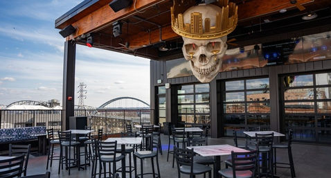 Rooftop patio bar with high-top tables and glass garage doors, a giant crowned skull chandelier overhead, and an arched-bridge city skyline view.