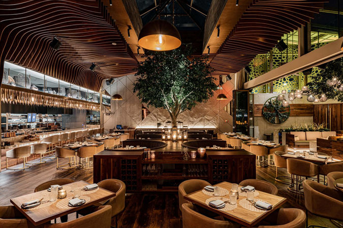 Upscale modern restaurant interior with a central indoor tree beneath sculpted wooden wave ceiling, warm pendant lighting, leather chairs and wooden tables set for dining, open kitchen and cozy banquettes