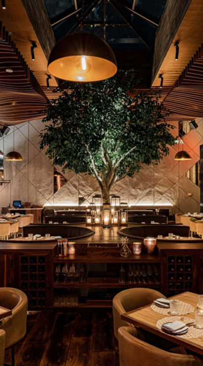 Upscale modern restaurant interior with a central indoor tree beneath sculpted wooden wave ceiling, warm pendant lighting, leather chairs and wooden tables set for dining, open kitchen and cozy banquettes