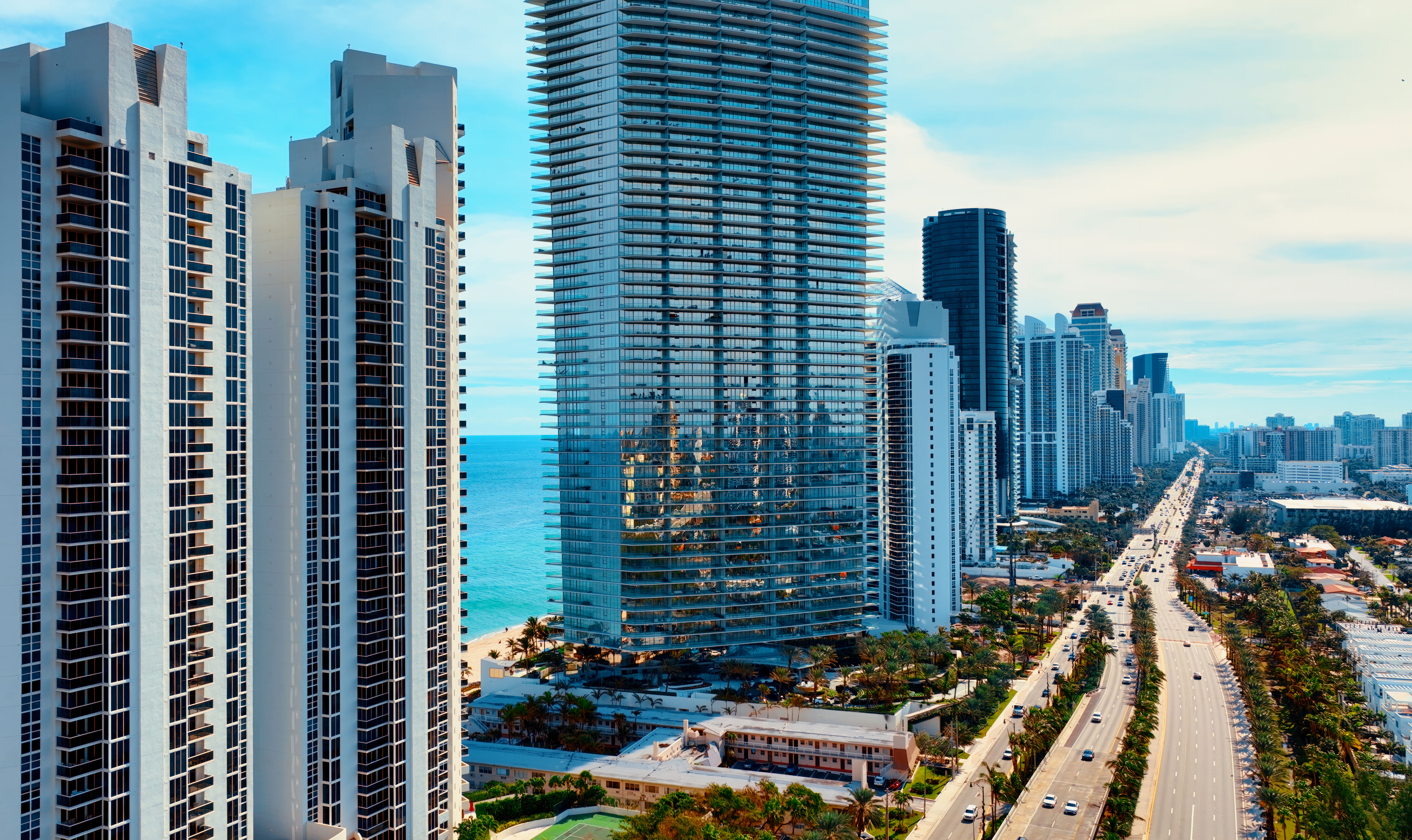 Sunlit aerial view of beachfront skyscrapers and a glass high-rise reflecting turquoise ocean, with a palm-lined multi-lane coastal highway stretching into the city skyline under a blue sky.