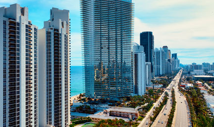 Sunlit aerial view of beachfront skyscrapers and a glass high-rise reflecting turquoise ocean, with a palm-lined multi-lane coastal highway stretching into the city skyline under a blue sky.