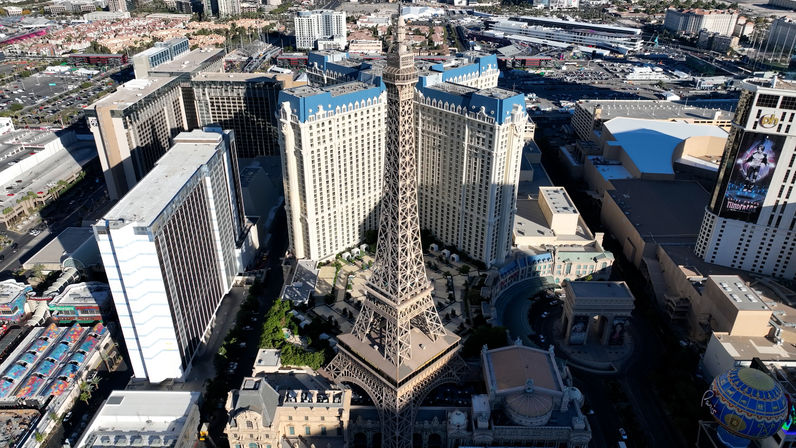 Aerial view of the Las Vegas Strip featuring an Eiffel Tower-style observation tower rising between sunlit high-rise hotels, pool courtyards, and busy city streets.