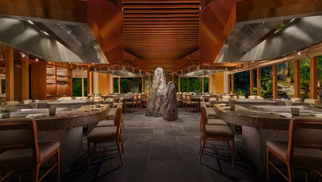 Sleek teppanyaki restaurant interior with curved granite grill counters, copper vent hoods, wooden slatted ceiling, central stone sculpture and garden views