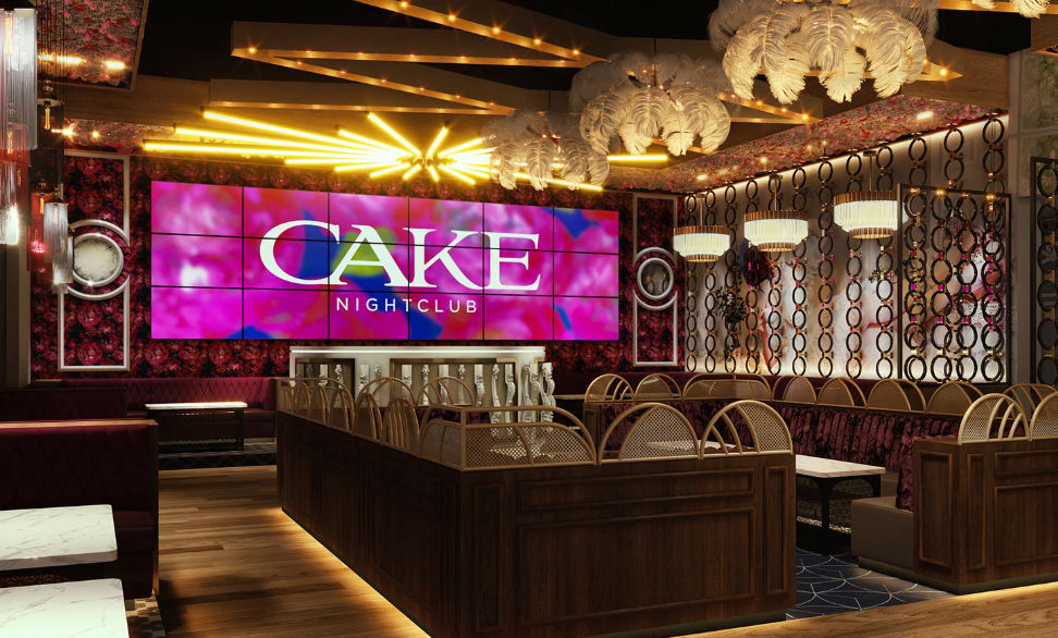 Upscale nightclub interior with vibrant pink LED video wall, plush velvet booths, wooden VIP banquettes, feather chandeliers and warm gold accent lighting.
