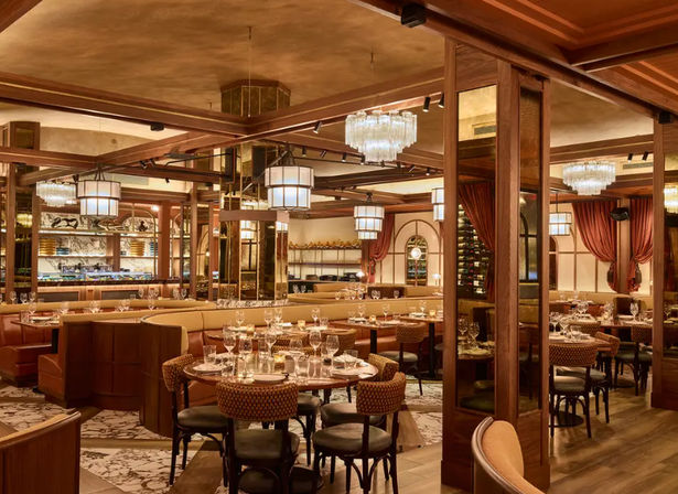 Upscale restaurant interior with warm wood paneling, plush banquette seating and round tables set with glassware and candles beneath crystal chandeliers — cozy, elegant dining space.