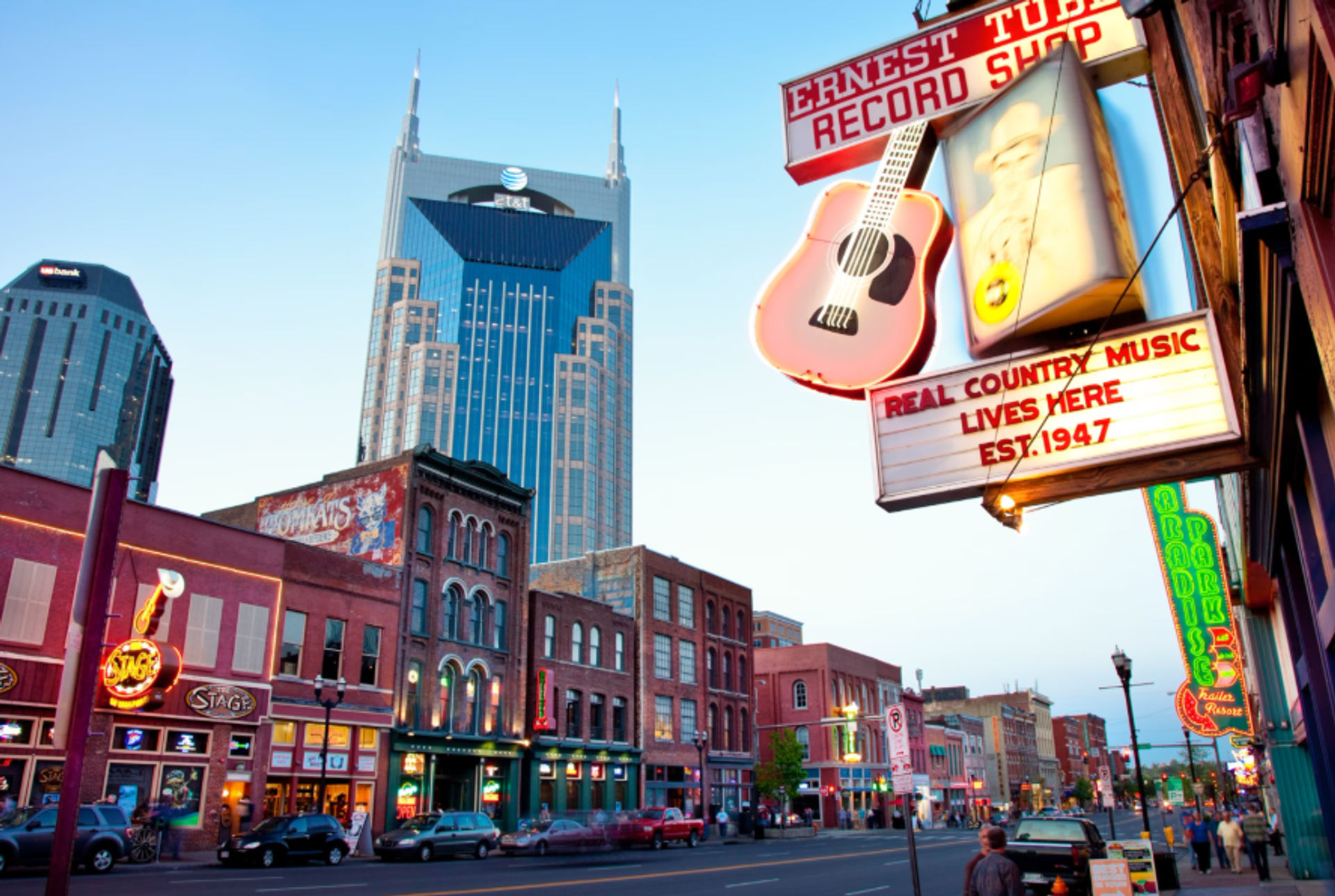 Lively Nashville Broadway at dusk with neon honky-tonk signs and an oversized guitar marquee, historic brick storefronts lining the street and a twin-spired skyscraper towering behind.