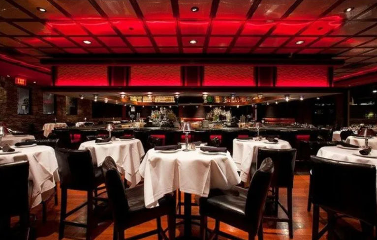 Upscale dining room with moody red ceiling lighting, white-tablecloth round tables set for service, black leather chairs, and a mirrored bar in the background.