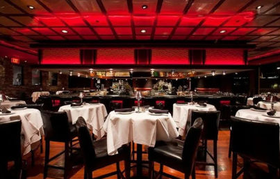 Upscale restaurant interior with red ambient ceiling lighting, round tables draped in white tablecloths set for dinner, black leather chairs and a bar with mirrored shelves in the background.