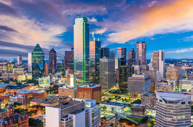 Aerial view of downtown Dallas skyline at sunset, vibrant colorful sky above illuminated glass skyscrapers, including a tall glass tower with green accent lighting and busy urban streets.