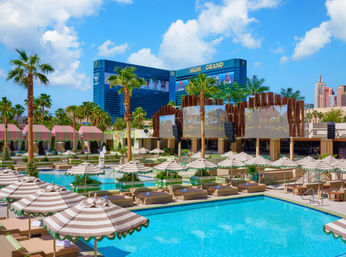 Sunny Las Vegas resort pool with turquoise water, striped umbrellas and daybeds, palm trees and cabana-lined deck with hotel towers in the background under a bright blue sky.