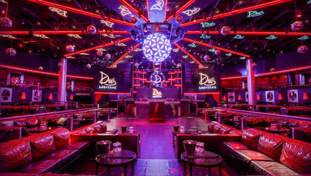 Vibrant Las Vegas nightclub interior with red and purple LED lighting, mirrored disco balls, a central DJ booth and leather VIP lounge seating with bottle service tables.