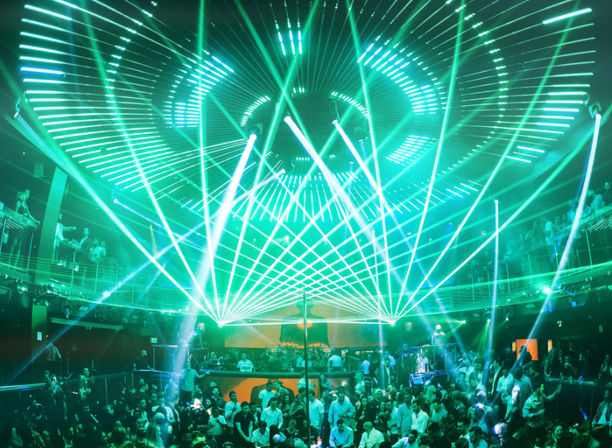 Packed nightclub dance floor with a lively crowd under swirling green laser beams and concentric LED ceiling lights, balconies and DJ booth visible