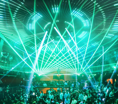 Packed nightclub dance floor with a lively crowd under swirling green laser beams and concentric LED ceiling lights, balconies and DJ booth visible