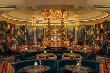 Opulent Art Deco hotel lounge and bar with towering golden palm chandeliers, curved green velvet banquettes, round tables, marble accents, tropical plants and warm ambient lighting