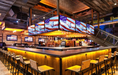 Rustic wood sports bar interior with a large illuminated central bar, rows of stools, mezzanine staircase, and multiple TVs showing American flag graphics.