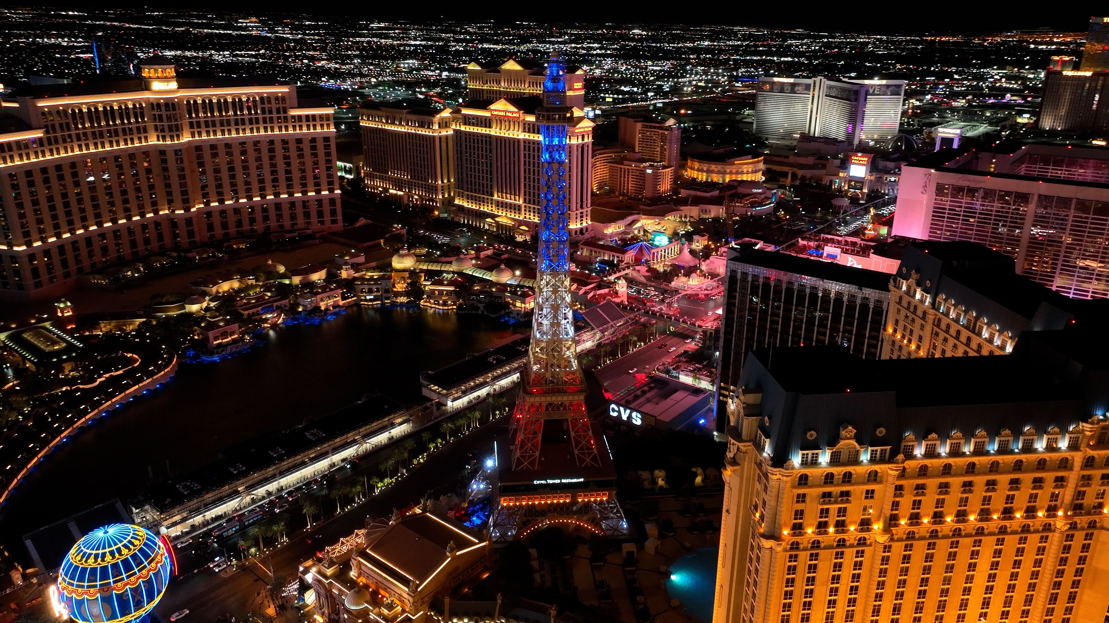 Aerial night view of the Las Vegas Strip featuring a lit Eiffel Tower replica, glowing hotel towers, neon signs and a reflecting lake of colorful lights.