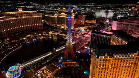 Aerial night view of the Las Vegas Strip featuring a lit Eiffel Tower replica, glowing hotel towers, neon signs and a reflecting lake of colorful lights.