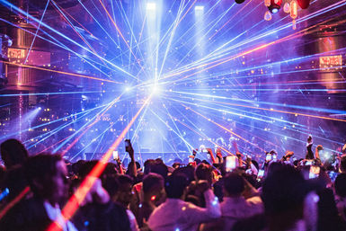 Packed indoor music venue crowd filming with phones as vibrant blue, purple and red lasers explode from the stage in an electric nightclub light show