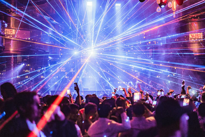 Packed indoor music venue crowd filming with phones as vibrant blue, purple and red lasers explode from the stage in an electric nightclub light show