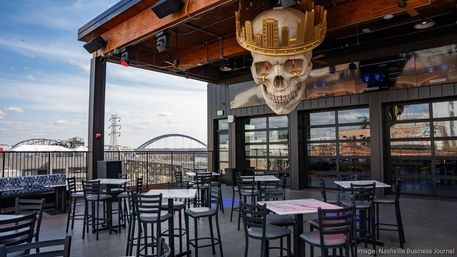 Nashville rooftop patio with empty high-top tables and chairs, giant gold‑crowned skull sculpture overhead and arched bridges over the river in the background.