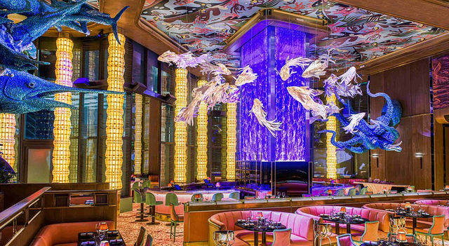 Vibrant upscale restaurant interior with aquatic-themed decor: suspended glass fish and octopus sculptures around a purple LED waterfall column, circular pink banquettes, tall golden column lights, and polished dining tables.