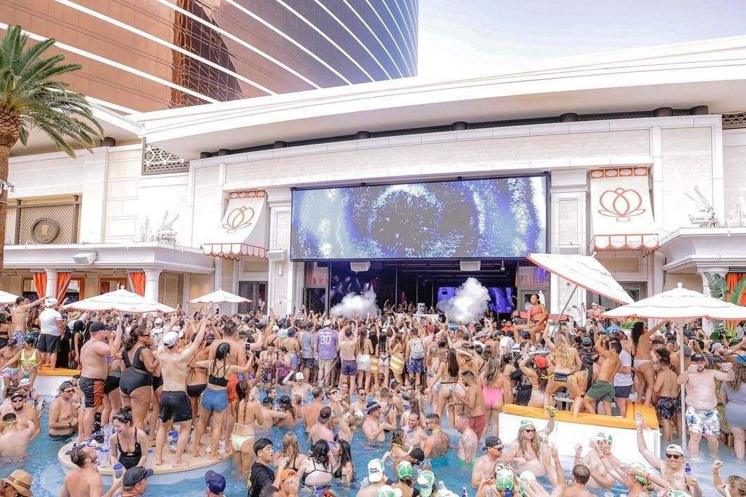 Energetic daytime resort pool party with hundreds in swimsuits dancing in and around a large pool, DJ on stage with giant LED screen, smoke effects, cabanas and umbrellas.