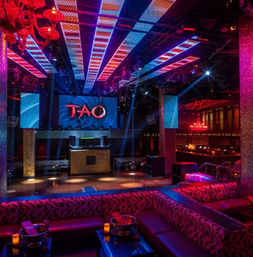 Vibrant nightclub interior with colorful LED ceiling strips, central DJ booth and dance floor, plush sectional lounges and low tables in a glowing VIP lounge.