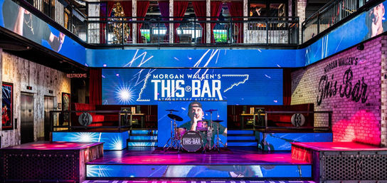 Vibrant two-level music bar interior ready for a live show: large LED stage screens, drum kit on an illuminated platform, mezzanine booths, exposed brick walls and neon restroom sign