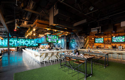 Spacious modern bar interior with illuminated central island bar and white stools, multiple LED wall displays, high-top communal tables on artificial turf, tropical-pattern accent walls and exposed industrial ceiling.