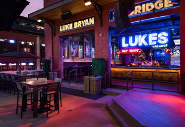 Vibrant neon-lit downtown patio and live-music bar at dusk, with high-top tables, outdoor stage, and colorful marquee signage.