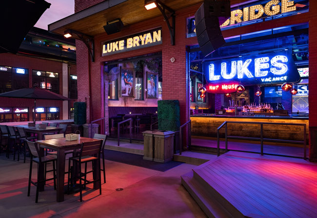 Vibrant neon-lit downtown patio and live-music bar at dusk, with high-top tables, outdoor stage, and colorful marquee signage.