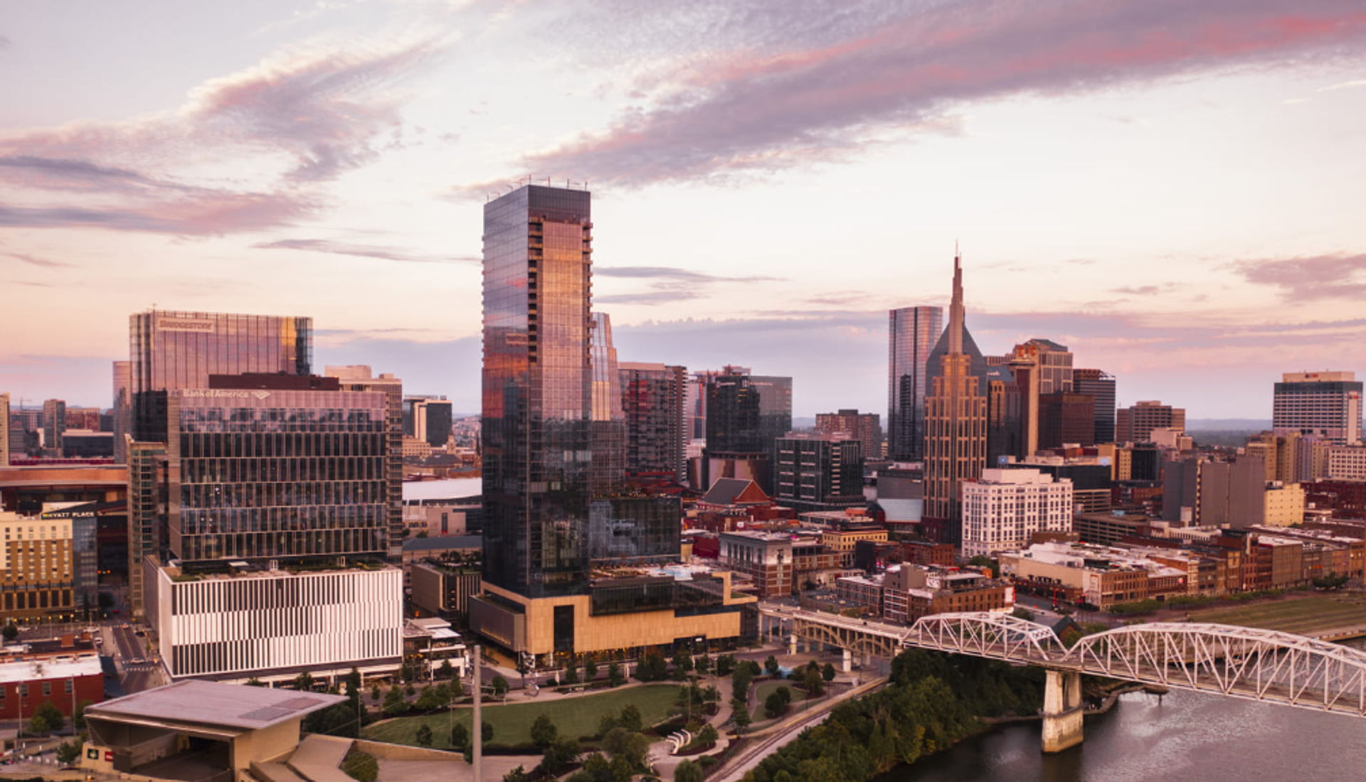 Downtown Nashville skyline at sunset with glistening glass skyscrapers, a white steel river bridge, and a pink-tinged sky over the riverfront.
