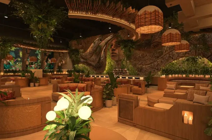 Cozy tropical-themed lounge interior with curved wooden booths, plush cushions, hanging wicker pendant lights, abundant indoor plants, candlelit tables, and a sculptural tree-face mural above the back bar.
