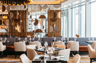 Chic modern restaurant interior with plush beige chairs, neatly set tables and wine glasses, a geometric metal room divider and floor-to-ceiling windows bathing the space in natural light.