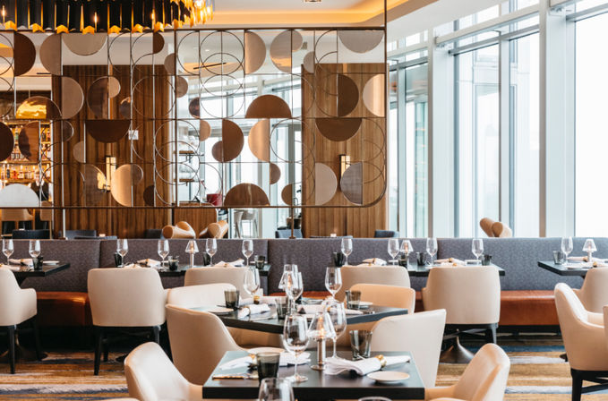 Chic modern restaurant interior with plush beige chairs, neatly set tables and wine glasses, a geometric metal room divider and floor-to-ceiling windows bathing the space in natural light.