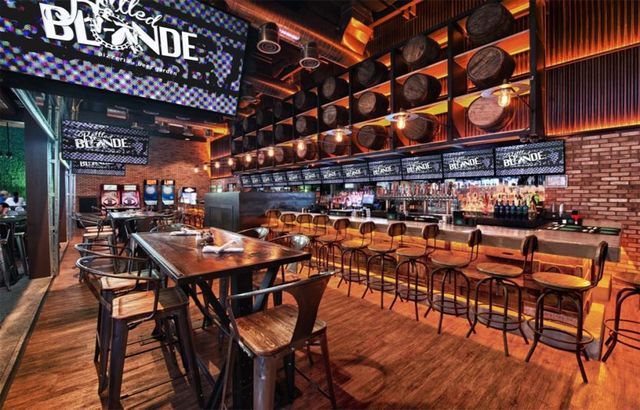 Modern industrial-style craft beer bar interior with a long wooden bar and metal stools, illuminated barrel wall, high-top tables, digital menu screens and warm ambient lighting.