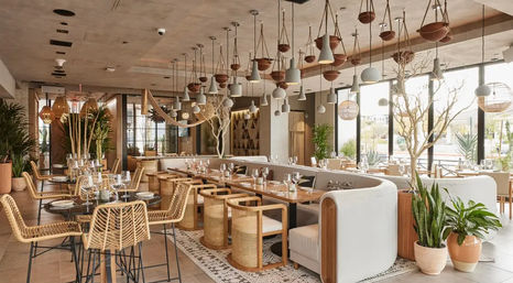 Boho-chic sunlit restaurant interior with rattan chairs, wooden tables, curved banquette seating, hanging pendant lights and abundant potted plants by floor-to-ceiling windows.