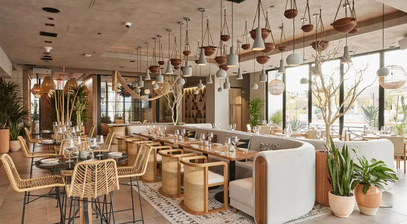Sunlit modern restaurant interior with natural wood tables, curved upholstered banquettes, woven rattan chairs, hanging pendant lights and potted plants by floor-to-ceiling windows.